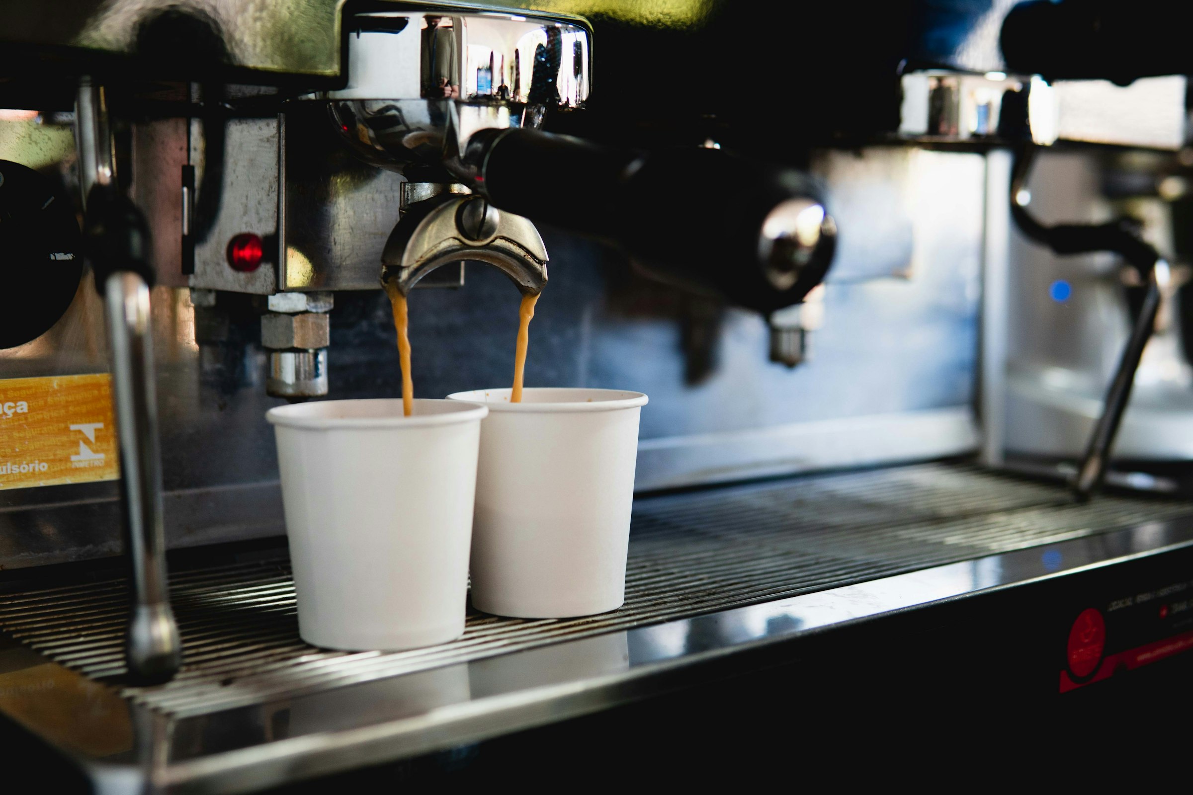 An espresso machine portafilter pouring two separate streams of coffee into two paper cups sitting side by side on a stainless steel drip tray — one source, two independent streams.