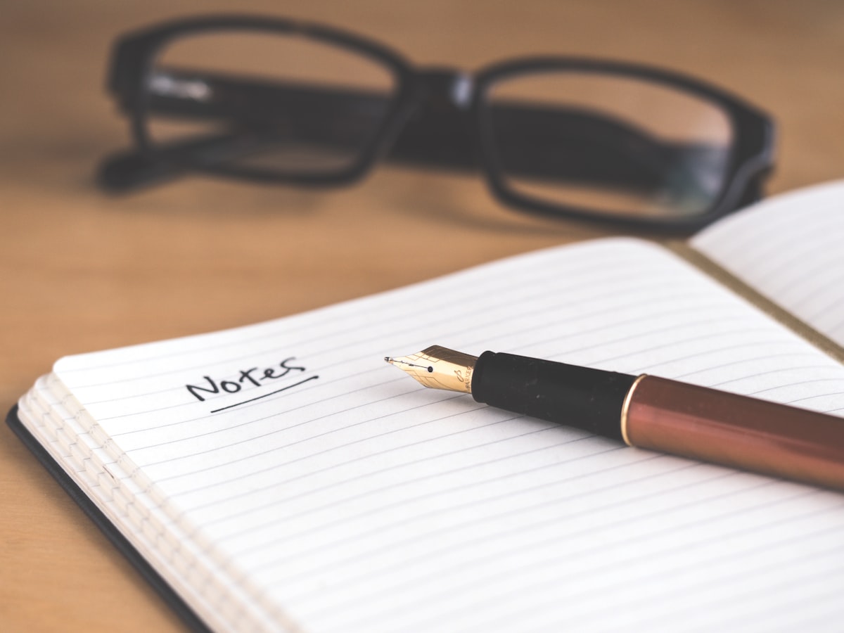 An open notebook with the word 'Notes' handwritten and underlined, a fountain pen resting on the page, reading glasses blurred behind it — the raw input a summarizer turns into one clean line.