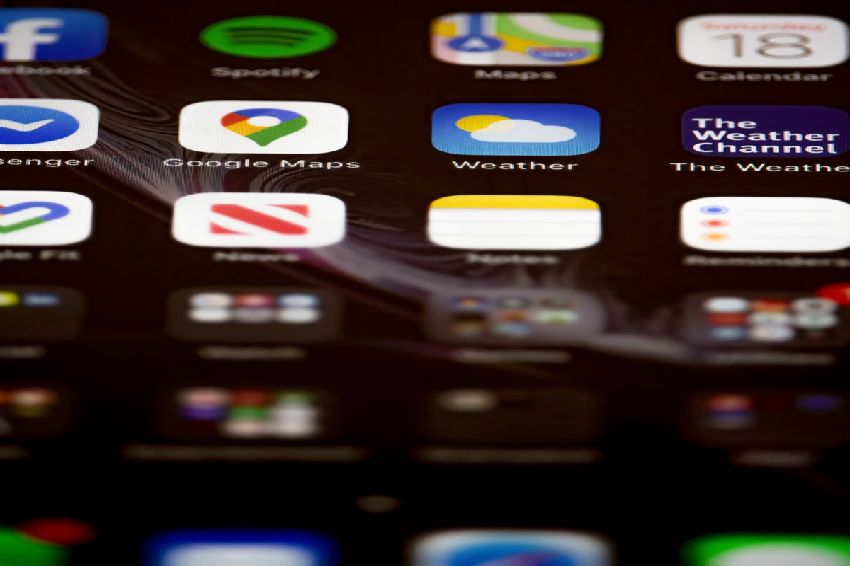 A close-up of an iPhone home screen — rows of app icons including Spotify, Maps, Weather, Google Maps, News, Notes — each a rounded-rectangle tile catching light slightly differently.