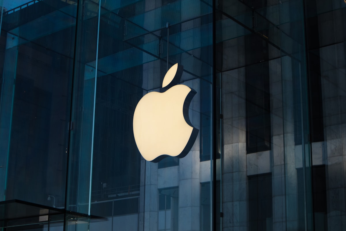 The glowing Apple logo on an Apple Store glass facade at night — representing Apple's expanding platform for third-party AI integrations through Siri Extensions