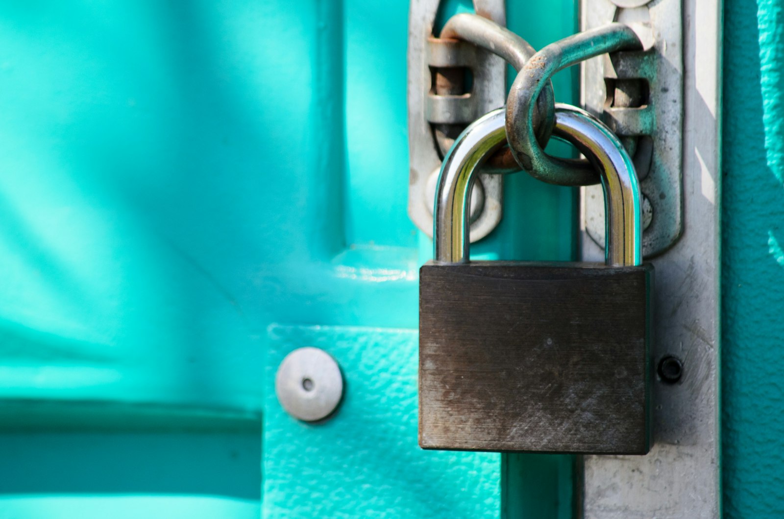 A heavy brass padlock fastened through the latch of a teal metal door.