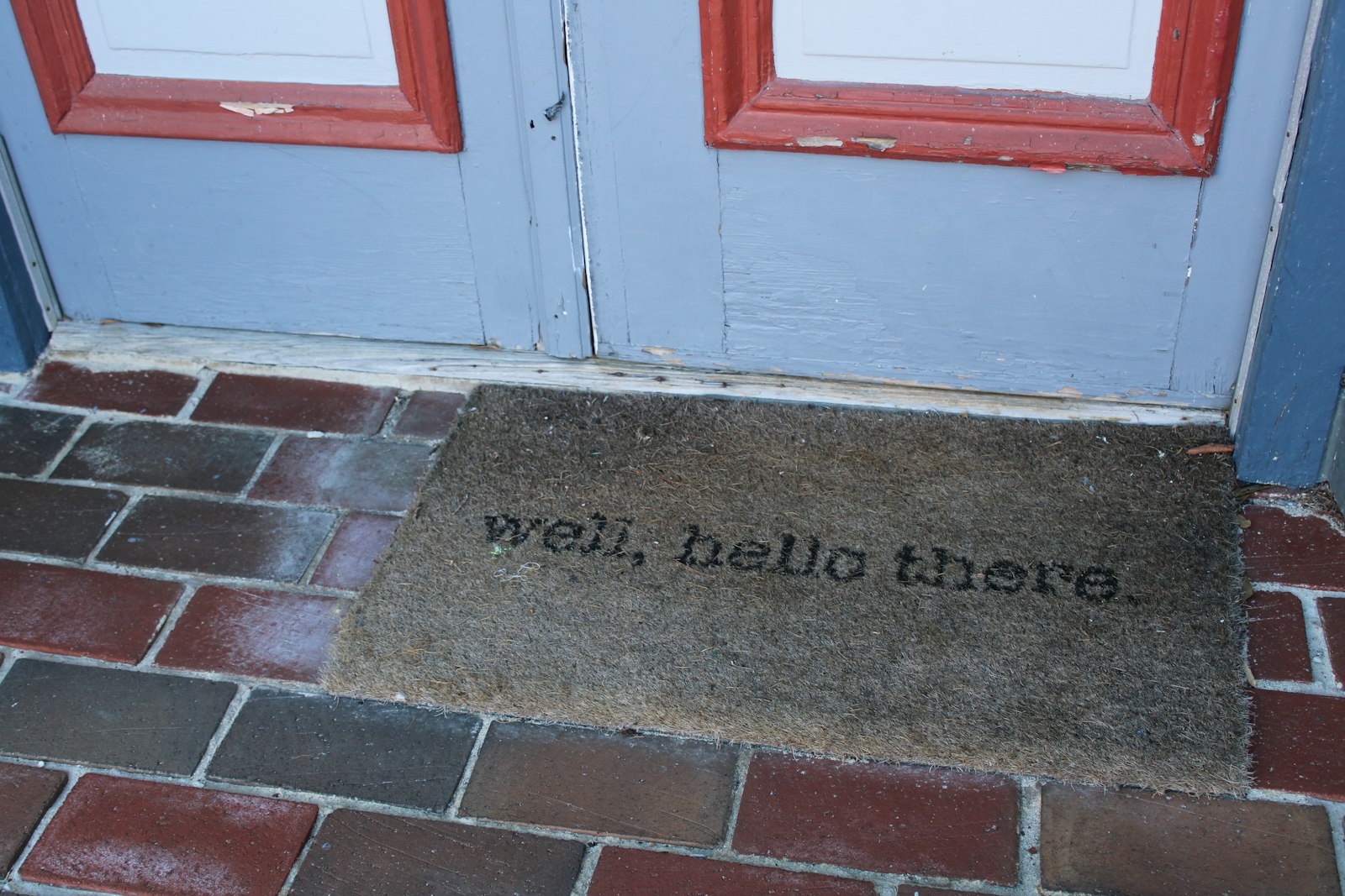 A coir doormat reading 'well, hello there.' on a red brick step in front of a blue door with red trim.