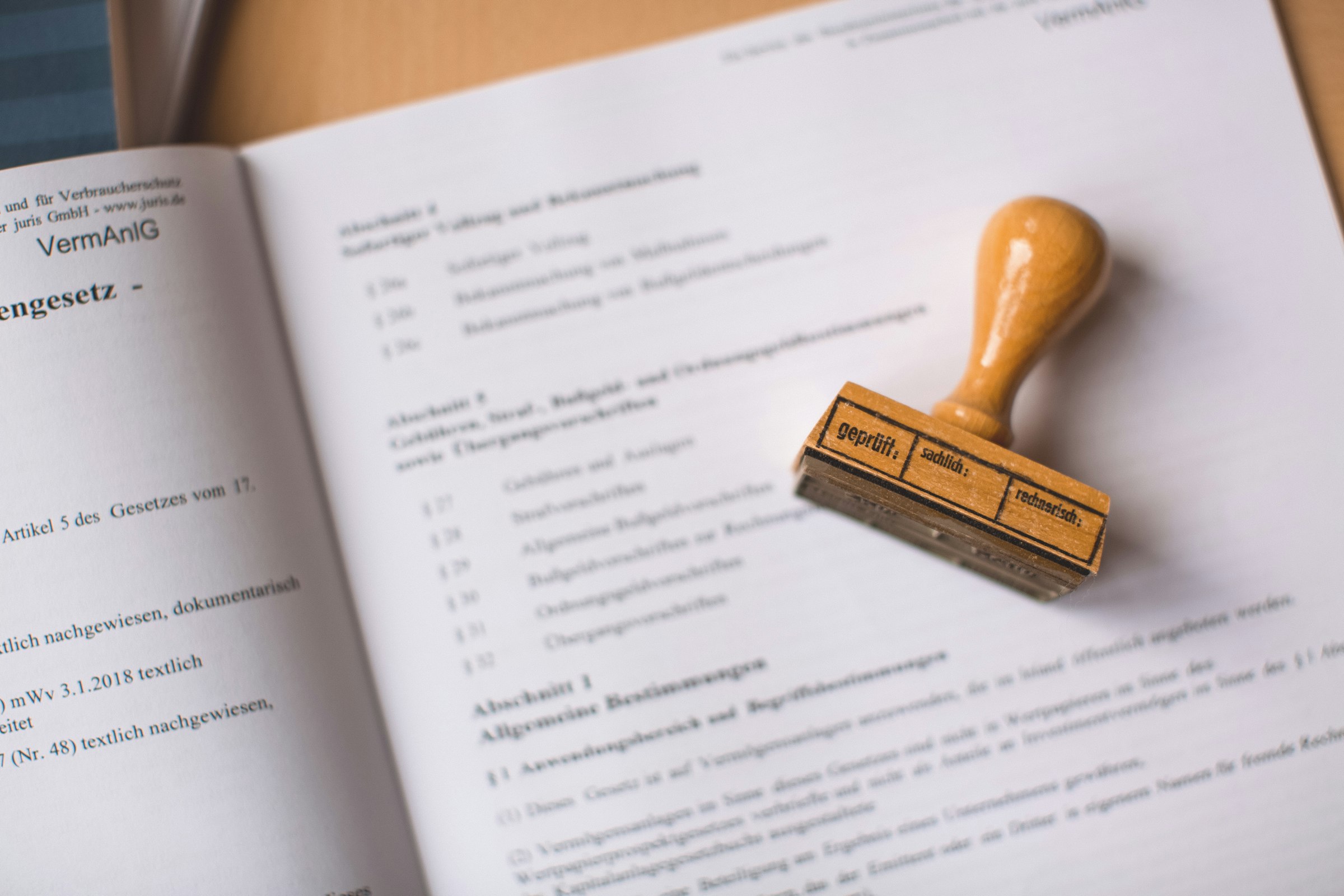 A wooden rubber stamp reading 'geprüft' (checked) resting on an open legal document, mid-stamp — something getting verified before it's allowed to count as approved.