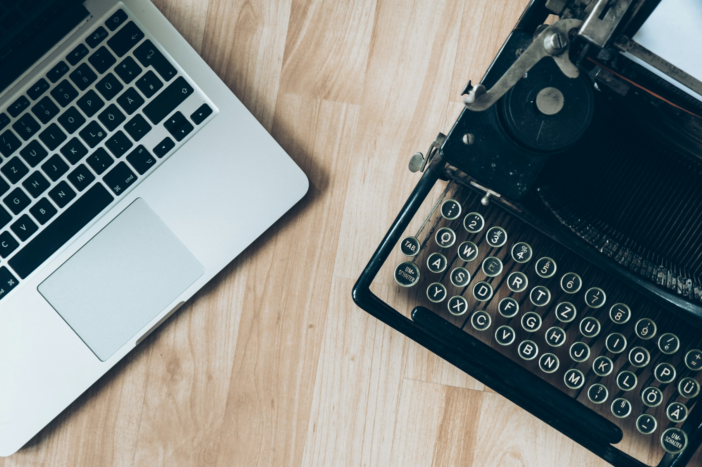 A vintage black typewriter and a modern MacBook sitting side by side on a wooden desk, shot from directly above — the old tool and the new one, both still doing real work.