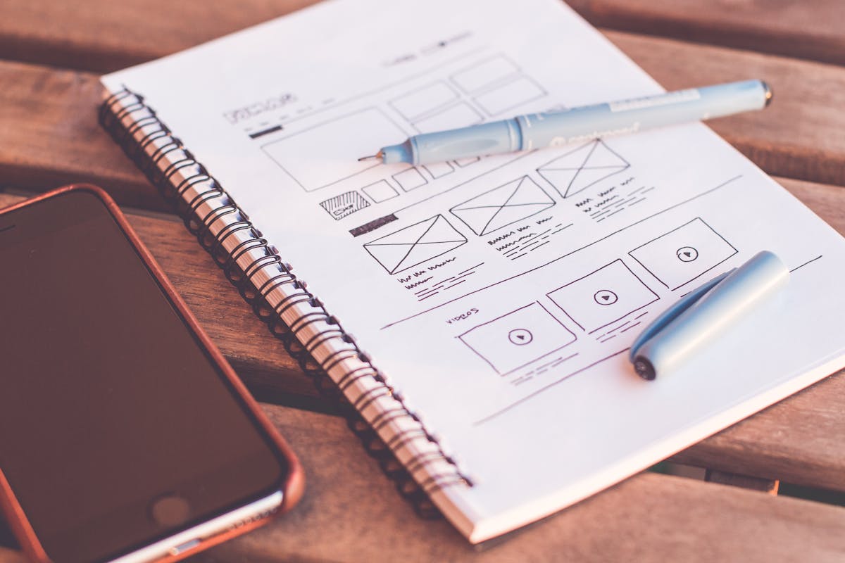 A notebook with hand-drawn wireframe sketches next to a smartphone and pen on a wooden table