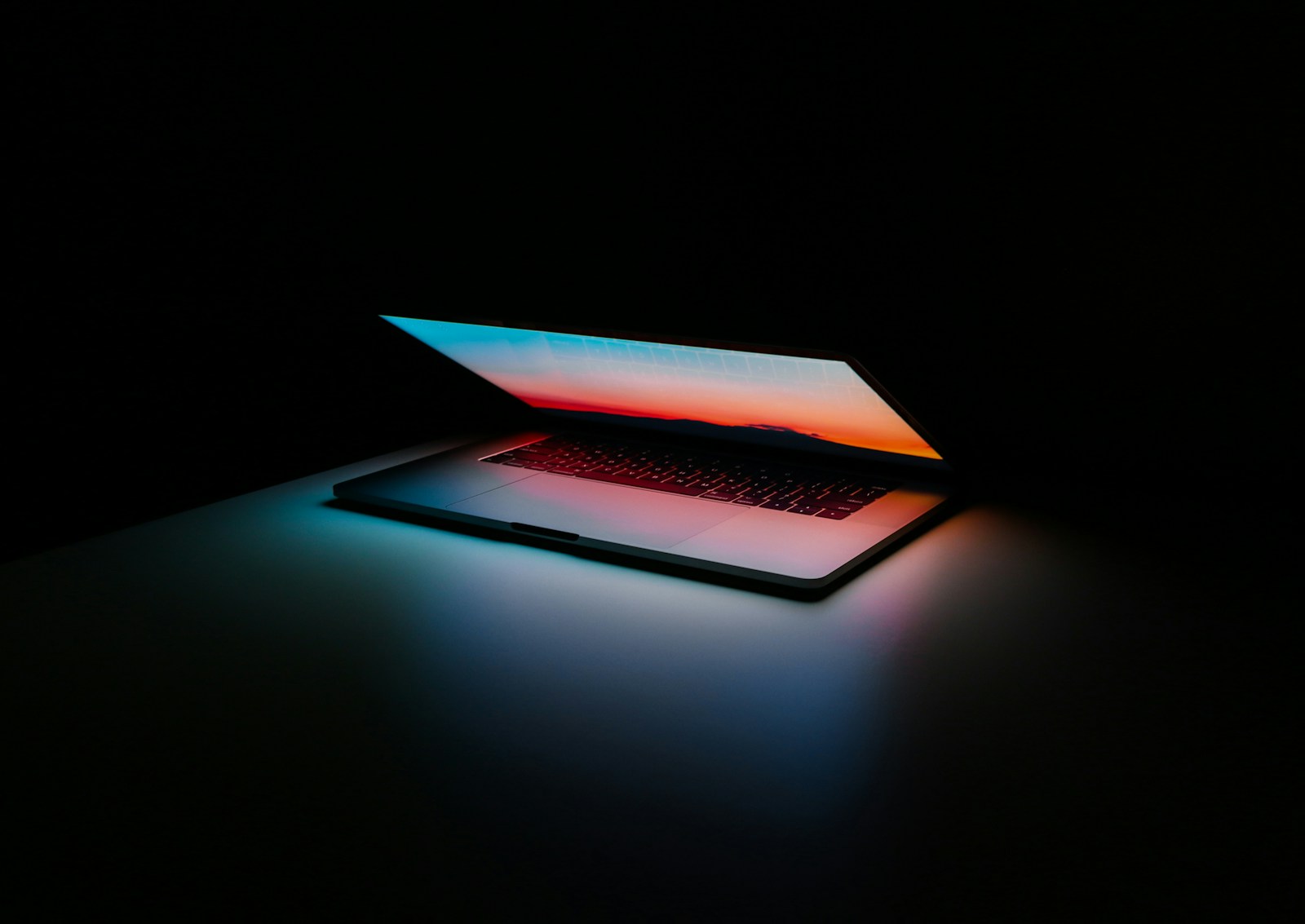 A laptop glowing in the dark, casting blue and orange light — reflecting Apple's 'All Systems Glow' WWDC 2026 tagline and the divided developer sentiment